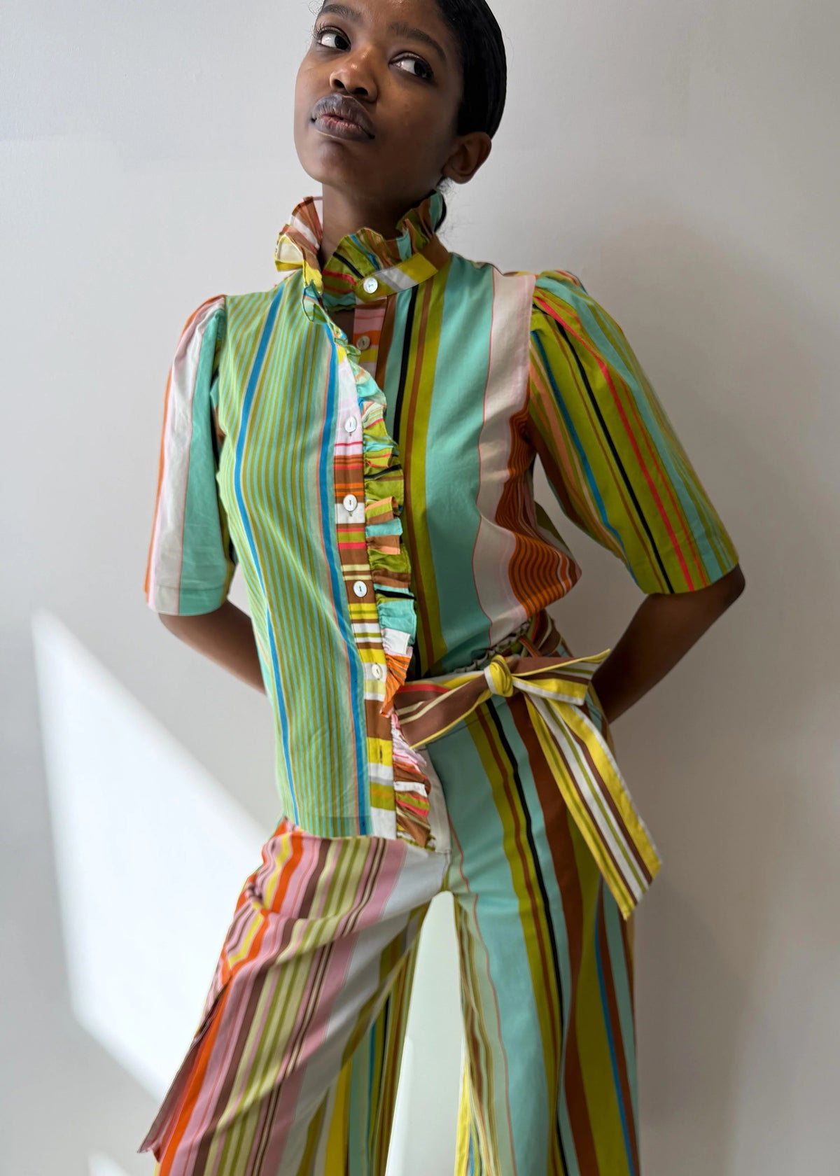 A person models the HOPE FOR FLOWERS Frilled Button Down Shirt in Combo Stripes by HOPE FOR FLOWERS, featuring vibrant vertical stripes and a ruffled collar, crafted from GOTS certified organic cotton for sustainability.
