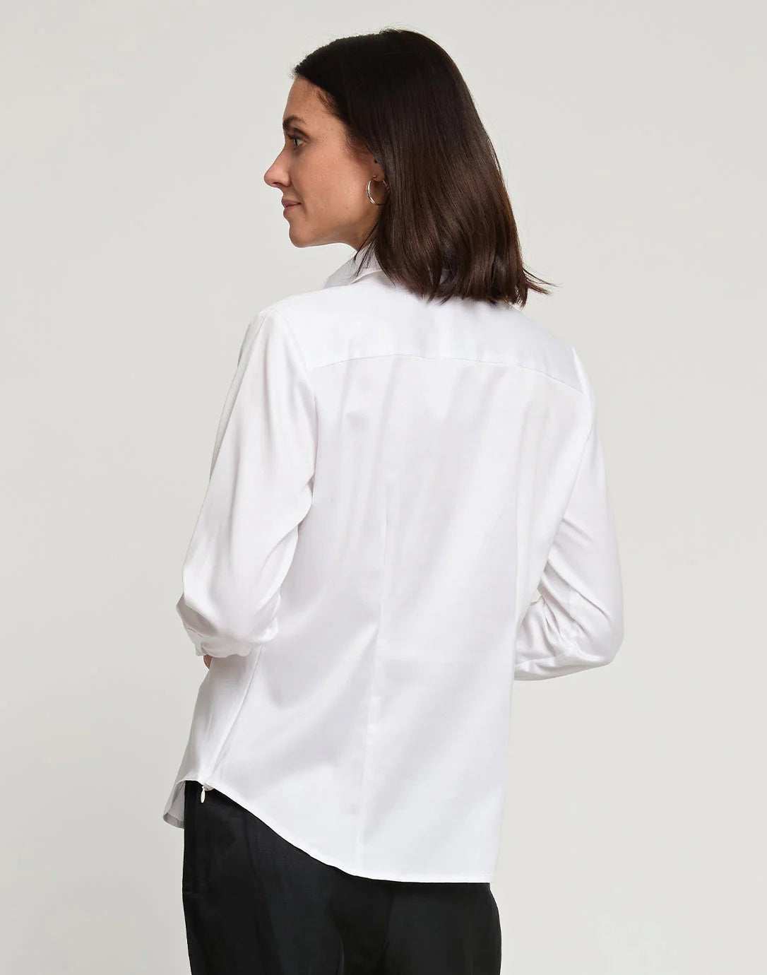 A woman with shoulder-length dark hair is shown from the back, wearing a white HINSON WU - MAYA Long Sleeve Cotton Satin Shirt and black pants. She is standing against a plain, light-colored background.
