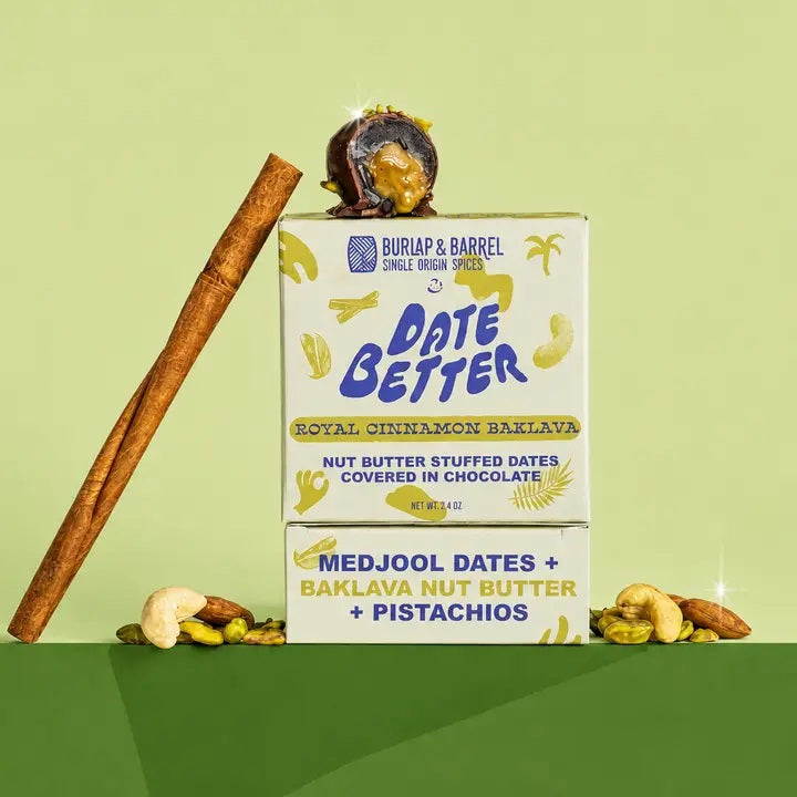 Two boxes of DATE BETTER - ROYAL CINNAMON BAKLAVA by DATE BETTER SNACKS, featuring nut butter-stuffed Medjool dates topped with chocolate, a cinnamon stick, cashews, and pistachios arranged against a green backdrop—the perfect gluten-free snack for any occasion.