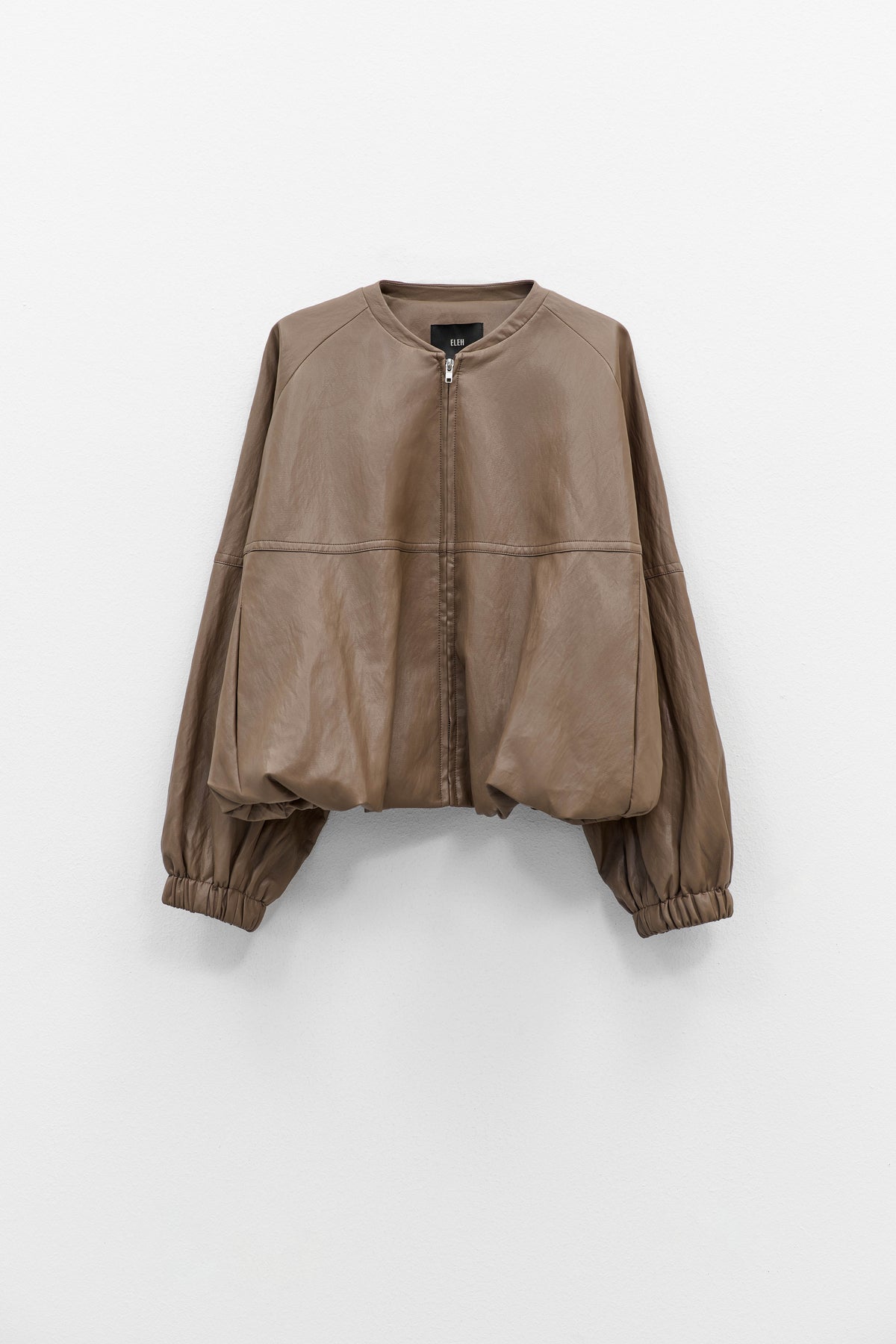 The ELEH - THE MOD BOMBER JACKET by ELEH is a brown leather jacket featuring a sculptural design, front zipper, round neckline, and long oversized sleeves with gathered cuffs, shown against a plain white background.