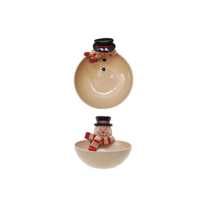 A cozy holiday table features the CREATIVE COOP STONEWARE BOWL WITH SNOWMAN HEAD, filled with treats and surrounded by snowman mugs and a teapot, all on a wooden table with a pink cloth.