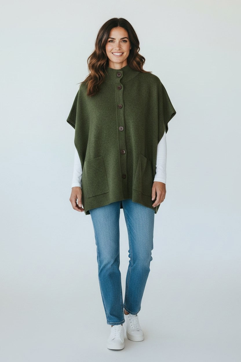 WOMAN wearing the KINROSS CASHMERE Short Sleeve Button Mock Cardigan in Highland layered over a white long-sleeve top and jeans, standing indoors near a large window and smiling softly at the camera. Her look is completed with this oversized sleeveless cardigan by KINROSS for added warmth and style.