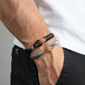 A close-up shows a wrist wearing MAKARLA’s MEN'S NAUTICAL ROPE BRACELET IN TOBACCO, a handmade adjustable accessory in black and tan, complementing a white shirt and dark pants.