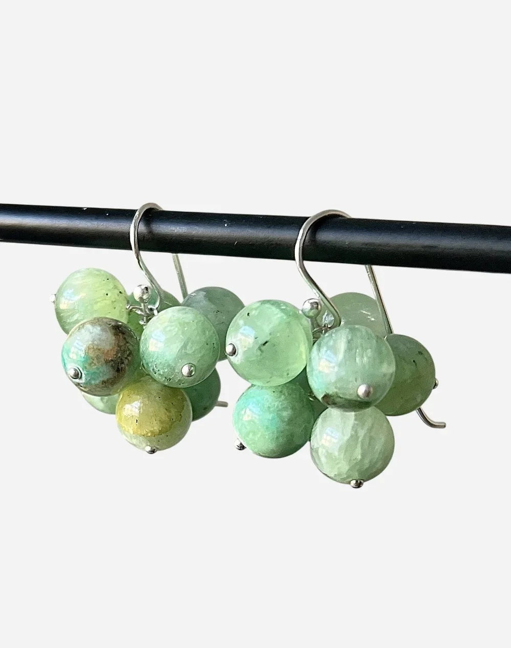 The ANN LIGHTFOOT - AZURITE JADE CLUSTER EARRINGS by ANN LIGHTFOOT feature clusters of round, green azurite jade gemstones in varying shades, suspended from sterling silver hooks and displayed on a black rod against a plain white background.