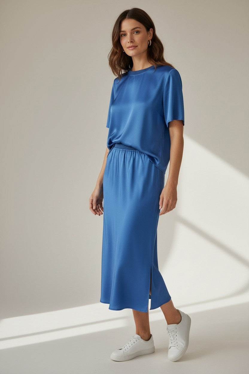 A woman with wavy brown hair smiles gently in a softly lit room, wearing the ROSE AND CROWN - SATIN TEE TOP and matching blue midi skirt with a side slit from ROSE & CROWN, paired with white sneakers.