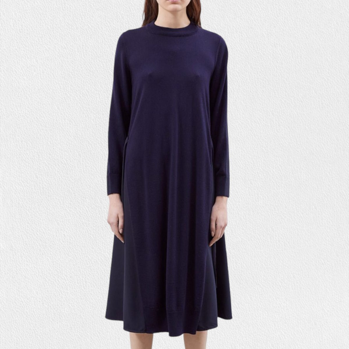 A woman models the MEIMEIJ - WOOL MIX MEDIA DRESS by MEIMEIJ, featuring long sleeves and a navy blue, knee-length wool design. She stands against a plain white textured background with her hair down and her hands relaxed at her sides.