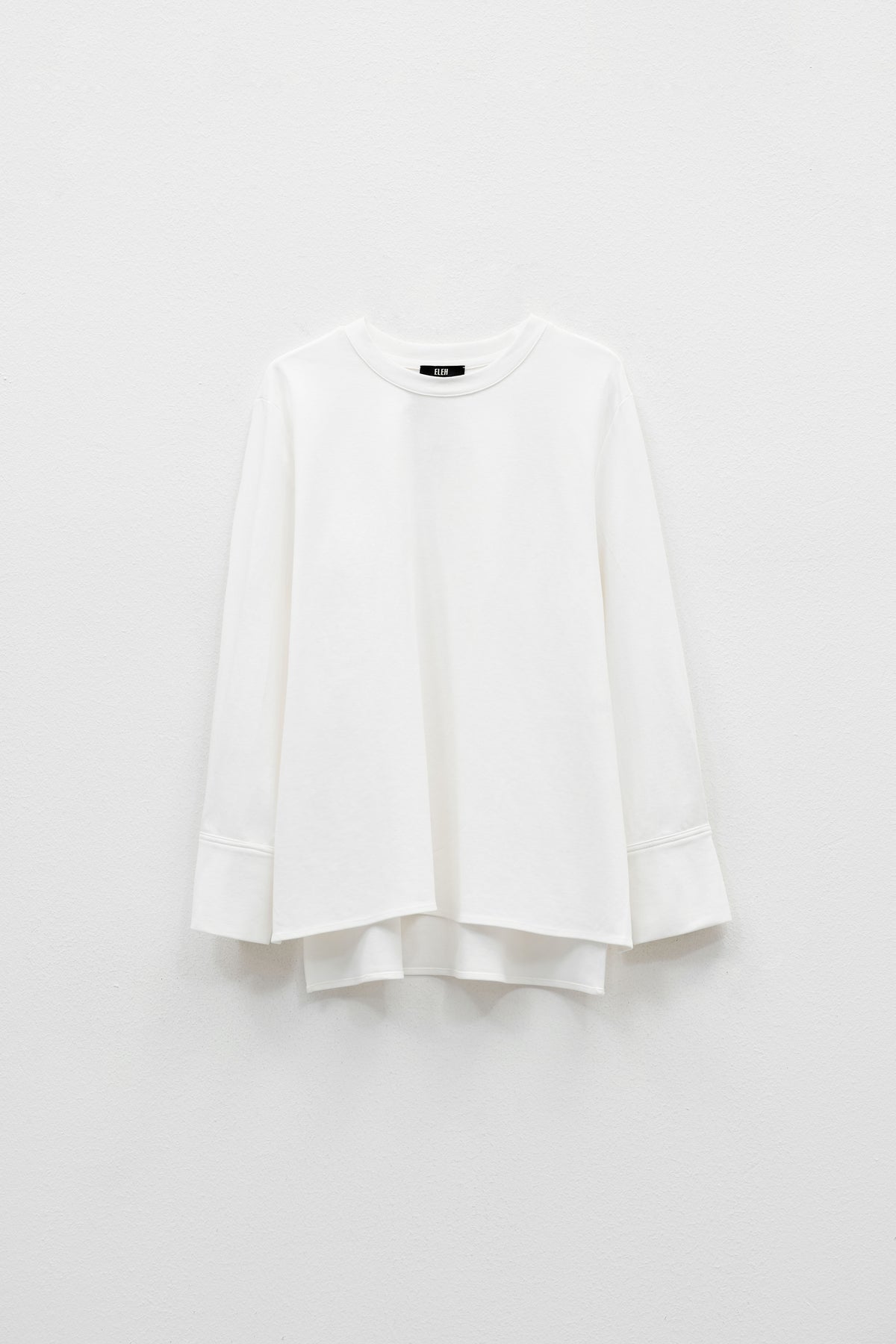 The ELEH - THE BIANCA CUFF TOP by ELEH is a plain white, long-sleeve crew neck shirt with wide sleeves, a loose fit, and an extended back hem for effortless style. Displayed against a white background, this versatile piece features a minimalist silhouette.
