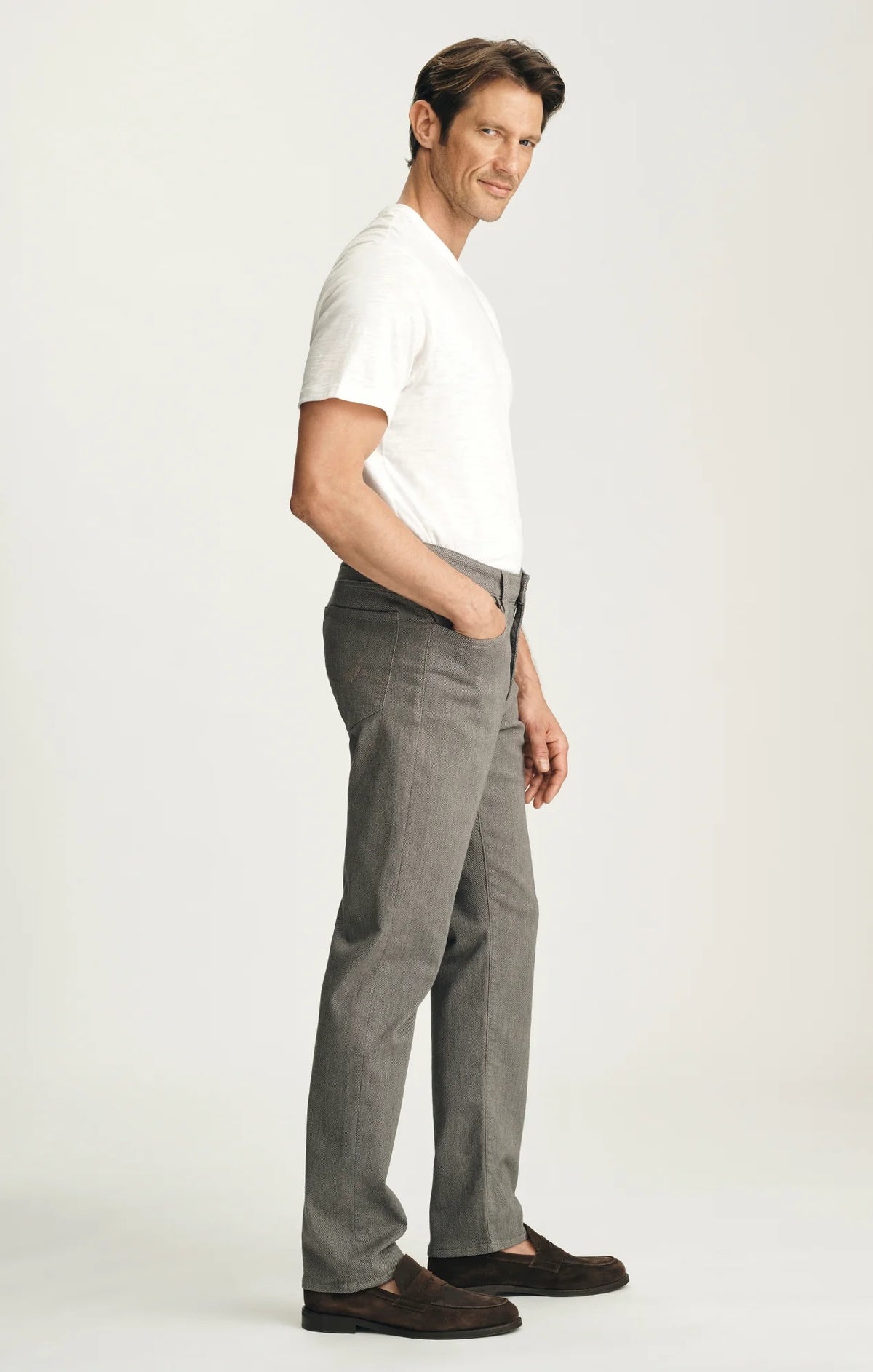 A man stands against a plain background wearing a dark brown sweater layered over a light collared shirt, paired with 34 HERITAGE - COURAGE STRAIGHT LEG PANTS IN STONE DIAGONAL by 34 HERITAGE, and brown shoes. One hand rests in his pocket and he maintains a neutral expression.