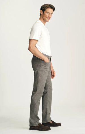 A man stands against a plain background wearing a dark brown sweater layered over a light collared shirt, paired with 34 HERITAGE - COURAGE STRAIGHT LEG PANTS IN STONE DIAGONAL by 34 HERITAGE, and brown shoes. One hand rests in his pocket and he maintains a neutral expression.