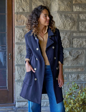A woman stands indoors by a window, smiling with her hands in the pockets of a FRANK & EILEEN Belfast Sweatshirt Trench Coat in Classic Navy from the California Heritage Collection. She wears a tan shirt, wide-leg blue jeans, and brown boots. A wooden stool is positioned behind her.