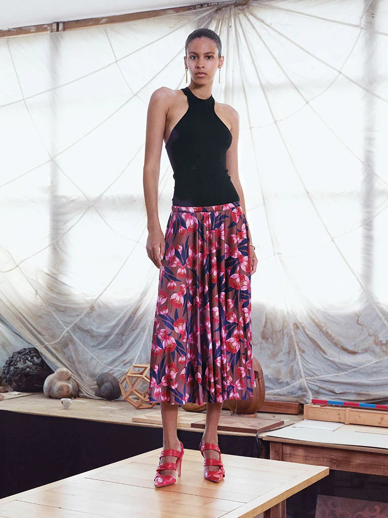 A woman with wavy hair smiles softly in a bright room, wearing a white V-neck blouse, brown strappy sandals, and the FRANCO FERRARI - FLORAL PRINT SKIRT—a high-waisted pink and navy made-in-Italy piece by FRANCO FERRARI.