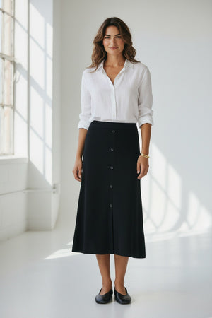 A woman with wavy brown hair stands in a sunlit room, smiling as sunlight streams through large windows. She wears a white button-up shirt paired with the HARTFORD - MELKA KNIT SKIRT IN BLACK by HARTFORD, black flats, and a gold bracelet.