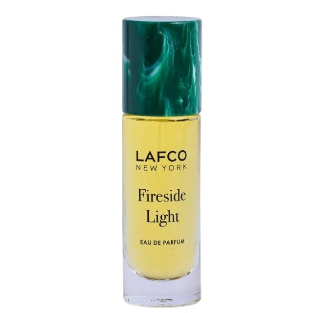 A 10ml bottle of LAFCO Fine Fragrance, Fireside Light rests atop a tree stump beside a vanilla bean, its green cap and brown smoky background evoking the warm woody notes of this vegan and cruelty-free scent.