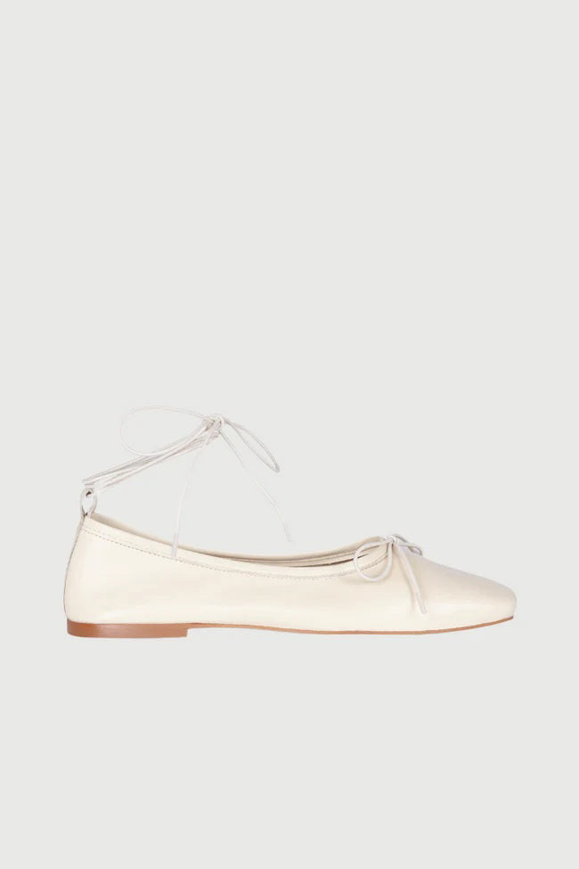 The Augusta - Sienna Ballet Flat by AUGUSTA is a single silver-colored shoe featuring a subtle metallic sheen and a simple bow on the front. It has a low brown sole and a pull tab at the back. Handcrafted in Spain from leather, this ballet flat is shown from the side against a light gray background.