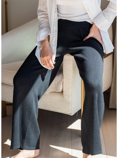 Seated barefoot on a light beige chair, a person wears MERSEA’s SEAPORT KNIT PULL ON PANT with side pockets, paired with a white top and open shirt. One hand rests in their pocket as sunlight streams onto their legs.