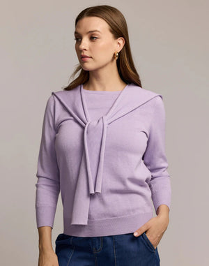 A woman with long brown hair and gold hoop earrings wears blue jeans and a light purple HINSON WU Sloane 3/4 Sleeve Tie Shoulder Cotton Silk Sweater, with a matching sweater draped over her shoulders and tied in front, looking to the side.