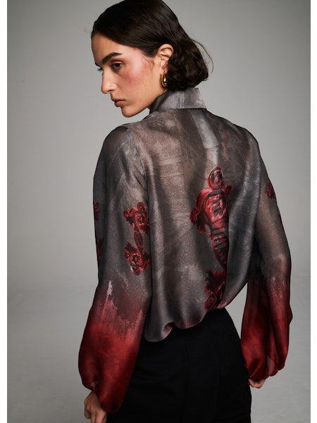 A person with short dark hair wears the MILVA MI - FLORAL RED BLOUSE by MILVA—featuring a gray and red floral pattern—and black high-waisted trousers, standing against a plain gray background.