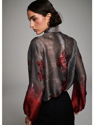 A person with short dark hair wears the MILVA MI - FLORAL RED BLOUSE by MILVA—featuring a gray and red floral pattern—and black high-waisted trousers, standing against a plain gray background.