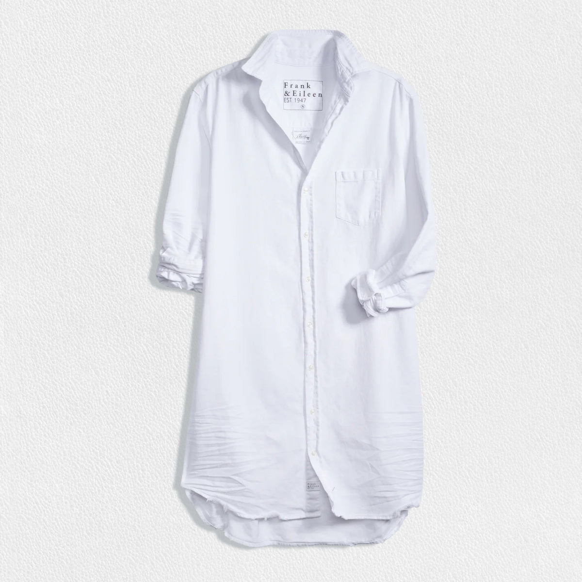 The FRANK AND EILEEN - MARY SHIRTDRESS IN TATTERED DENIM from FRANK &amp; EILEEN is showcased against a textured, off-white background. This long-sleeved dress boasts a collared neckline, a single chest pocket, and a slightly curved hemline. The sleeves are rolled up neatly, making it an ideal favorite for all four seasons.