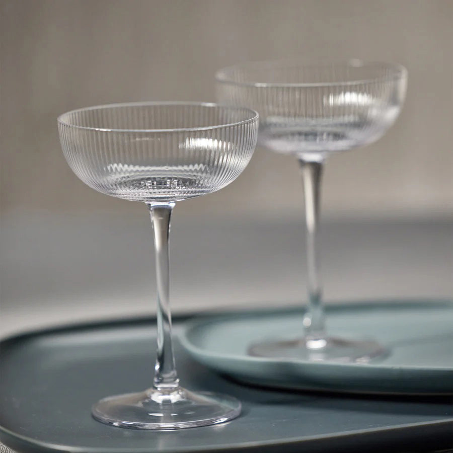 The OPTIC MARTINI GLASS from ZODAX is a clear, empty coupe glass with a capacity of 225 ml. It features a short stem and a wide, shallow bowl, adorned with subtle vertical ridges that add texture and elegance to its design. Set against a plain white background, its dimensions are perfectly showcased.
