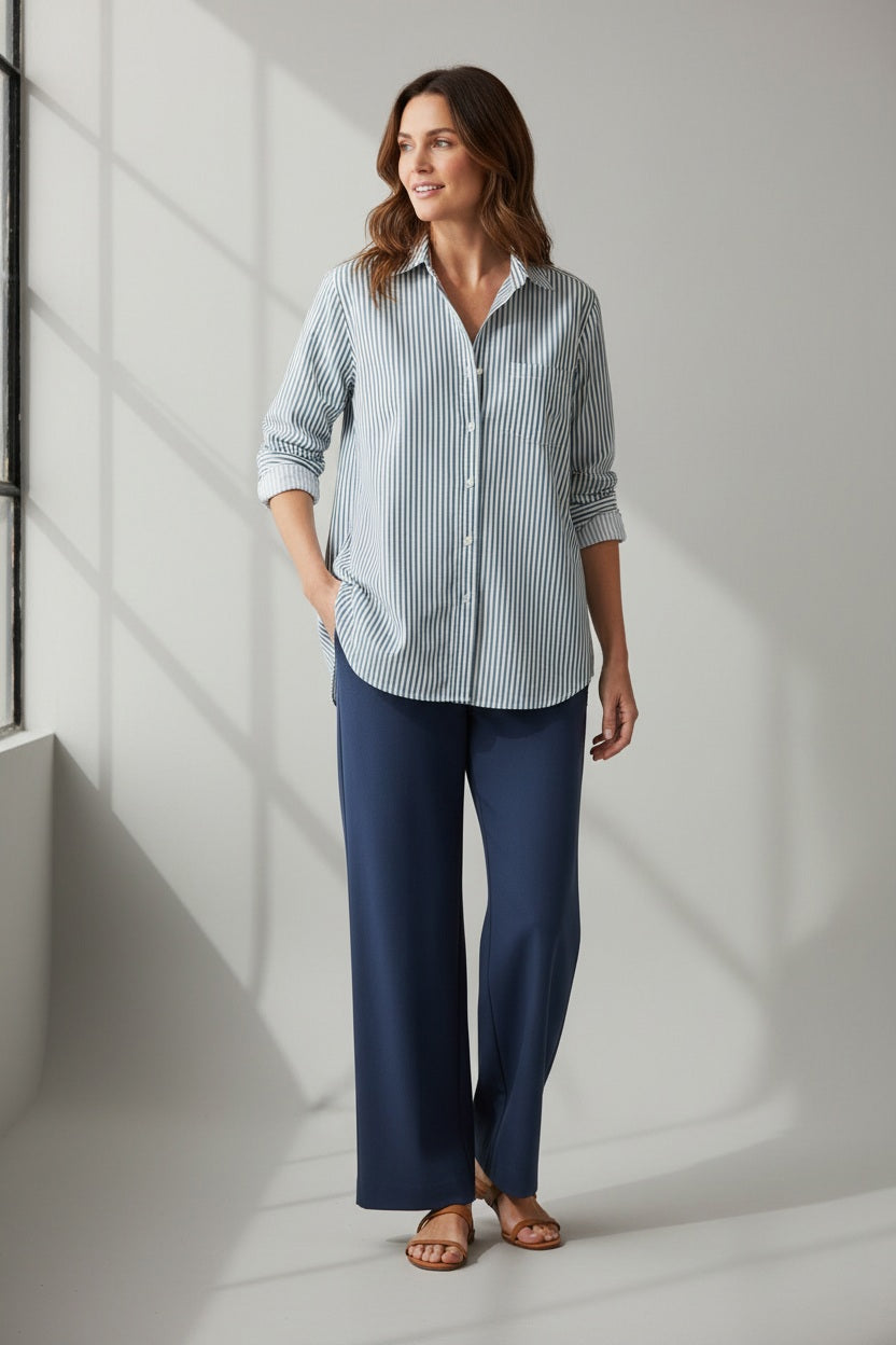 A woman stands indoors, smiling with one hand in her pocket, as sunlight streams through a window. She wears the FRANK & EILEEN EILEEN Relaxed Button Up Shirt in Italian Superluxe Cotton with green stripes, paired with navy wide-leg trousers and tan sandals. Her wavy brown hair frames her face.