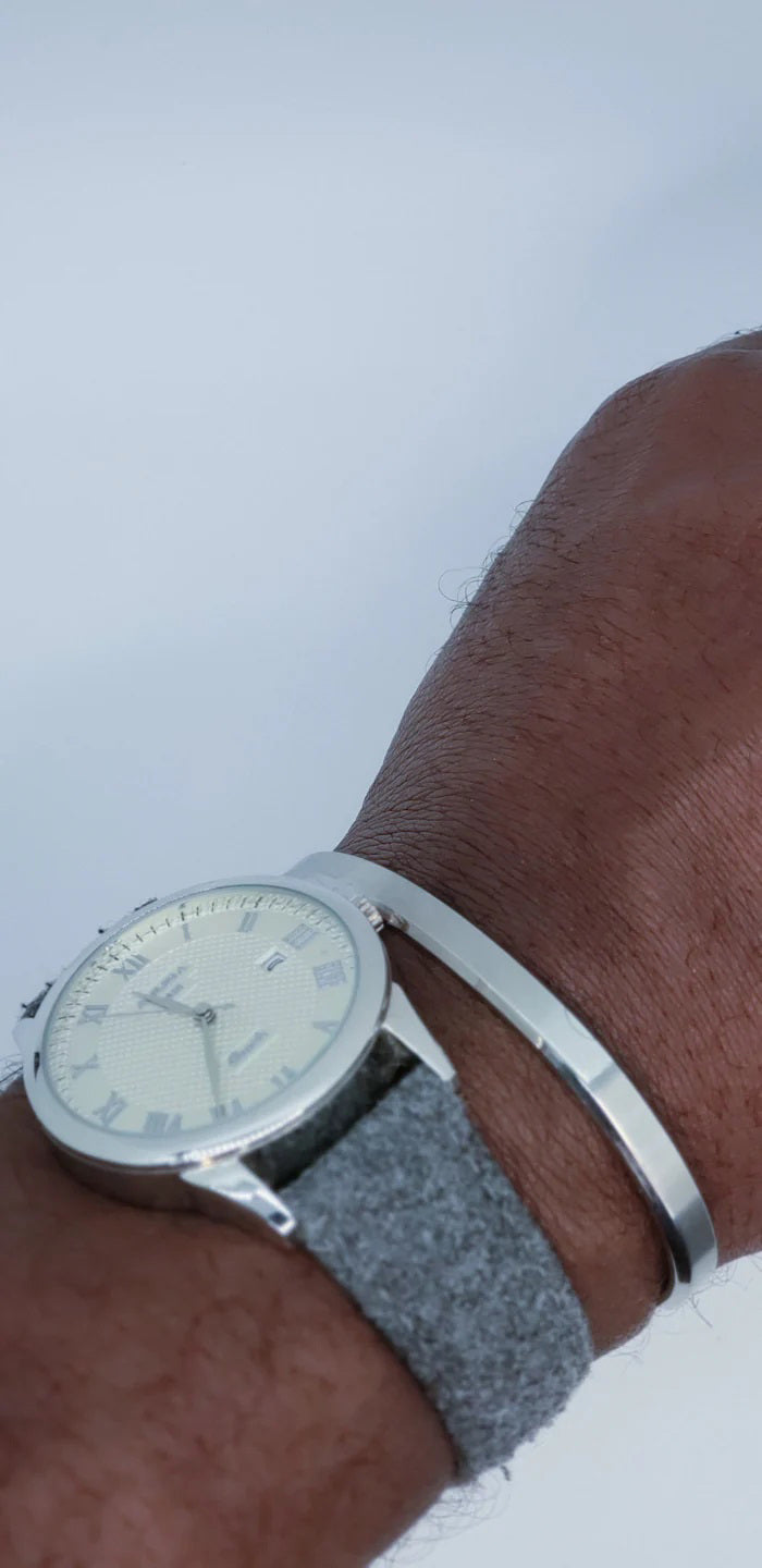 A close-up of a wrist wearing a silver analog watch with a textured gray band and the CLIFTON WILSON - SILVER STAINLESS STEEL BANGLE by CLIFTON WILSON, highlighting minimalist style against a plain white background.
