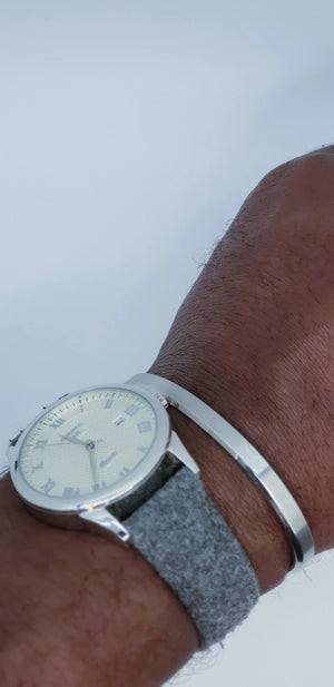 A close-up of a wrist wearing a silver analog watch with a textured gray band and the CLIFTON WILSON - SILVER STAINLESS STEEL BANGLE by CLIFTON WILSON, highlighting minimalist style against a plain white background.