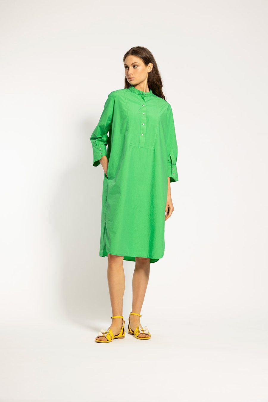 A woman stands against a plain background wearing the ACADEMIA Claude Shirt Dress/Long Tunic in a loose-fitting, bright green style with long sleeves and buttons down the front. She has her hands in her pockets and is looking to the side. Made from 100% cotton by ACADEMIA, she pairs the dress with yellow strappy sandals.