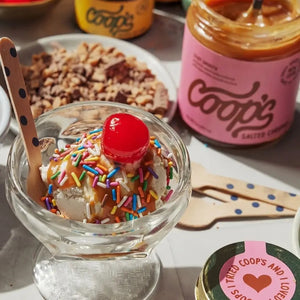 A glass bowl of ice cream topped with COOP’S - RAINBOW SPRINKLES, caramel sauce, and a cherry sits next to an open jar of Coop’s Salted Caramel Sauce and bowls of toppings on a table.