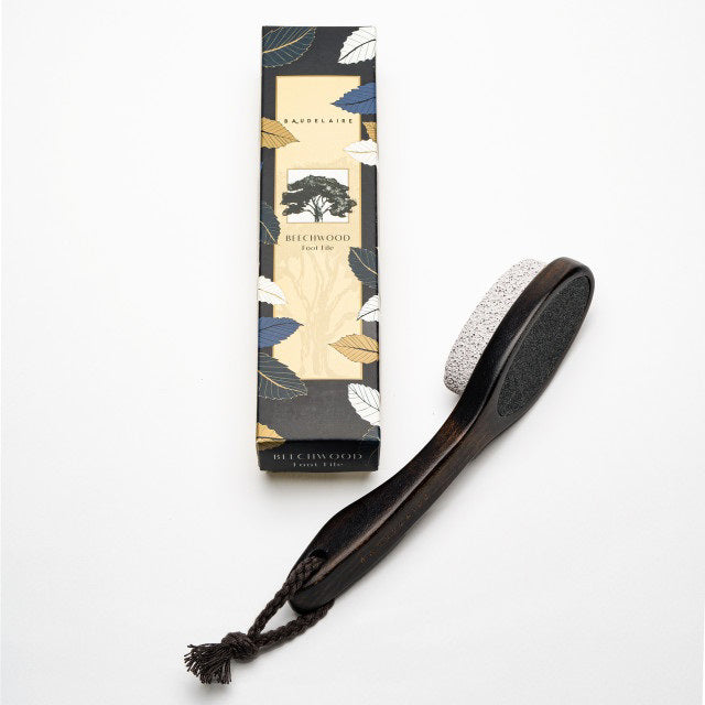 A two-sided foot file with a wooden handle and attached rope, featuring natural pumice on one side and a black abrasive surface on the other. The product, BAUDELAIRE - BEECHWOOD 8&quot; FOOT FILE by BAUDELAIRE, is laid next to its box, which has a botanical design and the branding &quot;Beechwood Foot File&quot; inspired by Japanese rituals.
