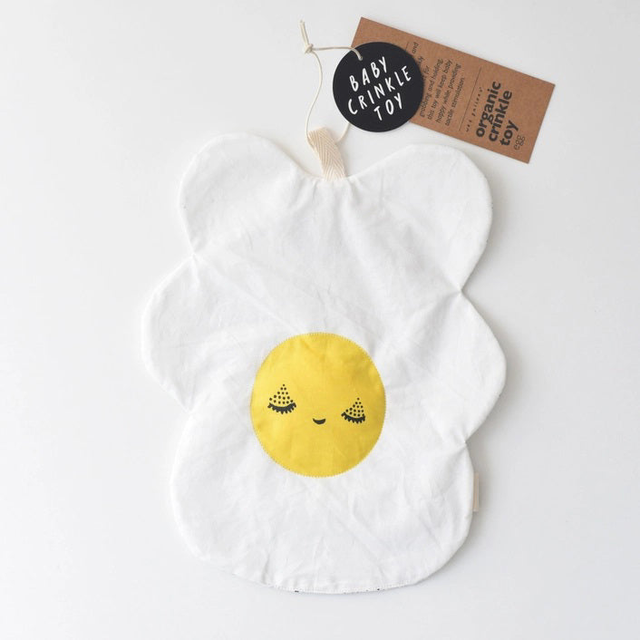 The WEE GALLERY - EGG CRINKLE TOY by WEE GALLERY is a white baby crinkle toy in the shape of a flower with a yellow circle at the center that features a smiling face with closed eyes. Made from organic cotton, it comes with a brown tag inscribed "Baby Crinkle Toy - Organic Crinkle Toy." The toy is displayed hanging on a white hook.