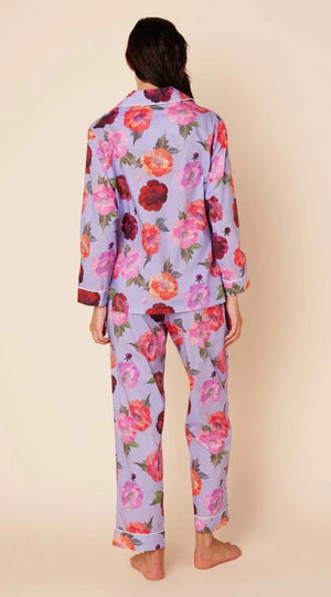 A woman is standing against a plain background, smiling downwards with her hands in the pockets of her Cat's Pajamas - Lavender Floral Luxe Pima Cotton Pajamas. The pajama set from Cat's Pajamas features a vibrant floral pattern with large red, pink, and purple flowers. She is barefoot and appears relaxed.
