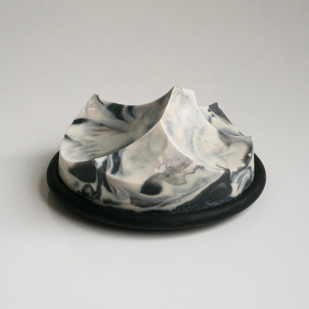 The UME STUDIO UME - JASMINE YLANG YLANG ERODE SOAP, a handcrafted black and white marbled bar shaped like a mountain range, rests on a round black dish against a plain, light background.