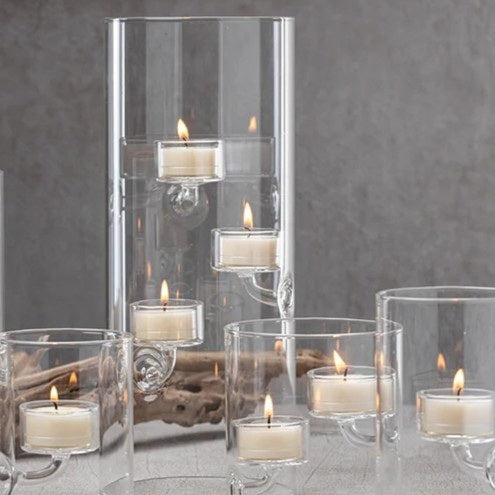 Five ZODAX SUSPENDED GLASS TEALIGHT HOLDER - EXTRA LARGE candle holders with floating tealights are arranged on a gray surface, with driftwood in the background. The windproof holders vary in height for an elegant display.
