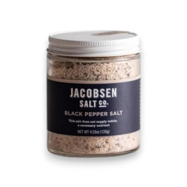 A small glass jar of JACOBSEN SALT CO. JACOBSEN SALT - INFUSED BLACK PEPPER SALT with a white lid rests on a light beige surface against a light gray background. The dark label with white text highlights this floral, spicy blend.