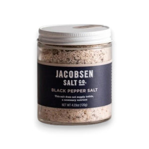 A small glass jar of JACOBSEN SALT CO. JACOBSEN SALT - INFUSED BLACK PEPPER SALT with a white lid rests on a light beige surface against a light gray background. The dark label with white text highlights this floral, spicy blend.