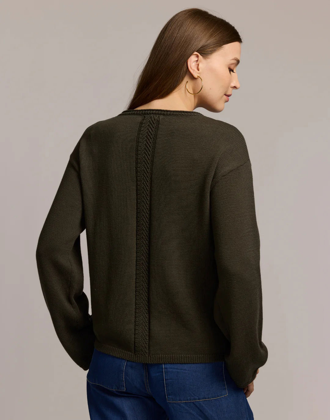 A woman with long brown hair and large hoop earrings models the HINSON WU AUDREY Long Sleeve Cotton Silk Sweater in tan with blue jeans, standing against a plain beige background, one hand raised near her shoulder.