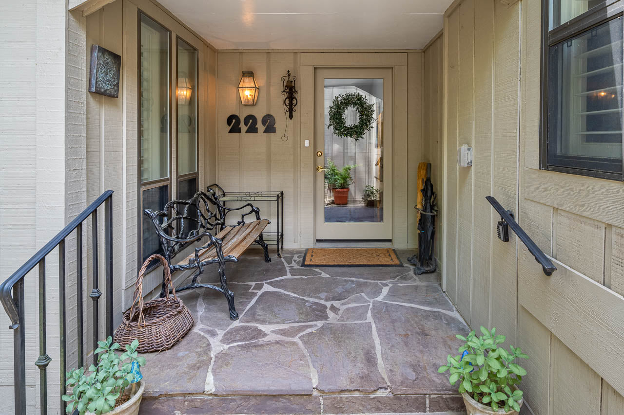 A welcoming front porch with stone flooring, a black bench, a wicker basket, two potted plants, wall-mounted lanterns, the house number 222, and a glass door decorated with a green wreath.
