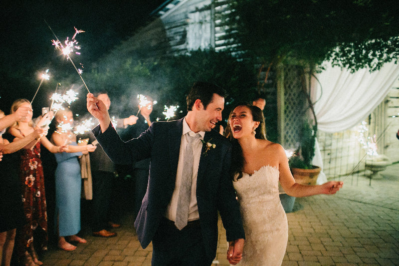 A bride and groom walk hand in hand at night, smiling and holding sparklers, surrounded by cheering guests celebrating outdoors.