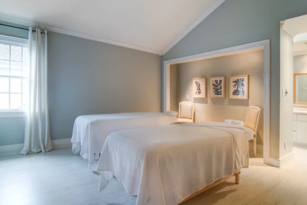 A serene spa room with two massage tables covered in white linens, light grey walls, three botanical prints on the wall, wicker chairs, and soft natural light from a window with white curtains.