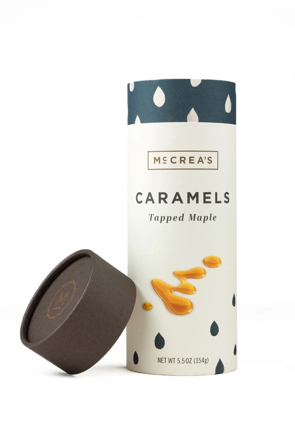 A cylindrical container of McCREA'S CANDIES Tapped Maple Syrup Caramels, featuring a dark brown lid, blue and white raindrop pattern, and caramel drizzle illustrations, inspired by New England maple syrup.