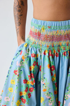 A person stands indoors wearing a white and blue striped vest and the HOPE FOR FLOWERS Bubble Hem Skirt in Chambray Floral by HOPE FOR FLOWERS. They have short curly hair and are wearing sandals.