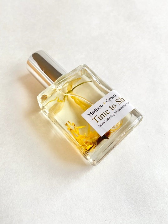 A clear glass bottle with a silver cap holds yellow, fast-absorbing aromatherapy oil infused with visible herbs and eucalyptus. The label reads “MADISON GREEN - TIME TO SHINE CONFIDENCE BOOST AROMATHERAPY BODY OIL MINI.”.