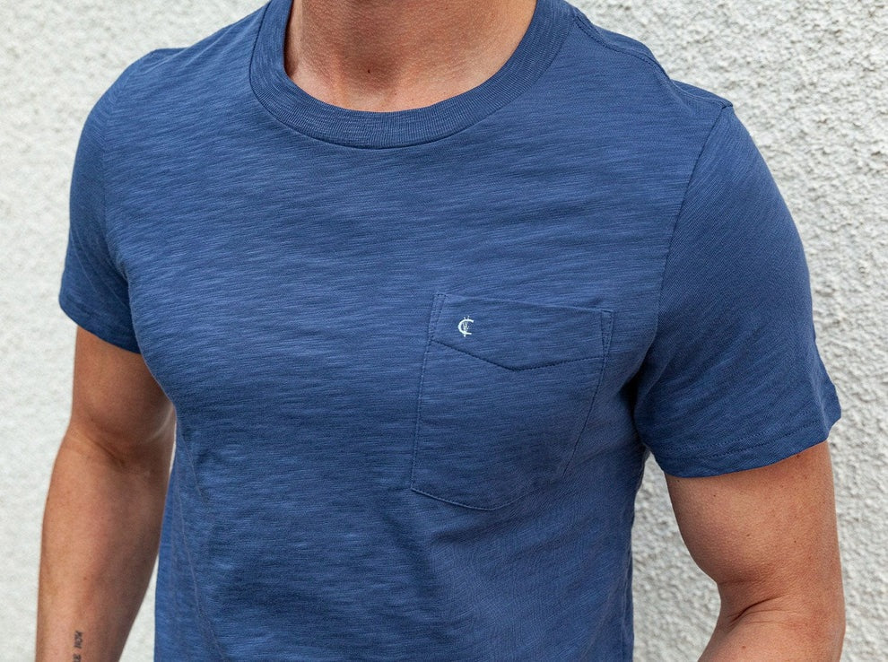 A man is wearing the CRIQUET Slub Cotton Short Sleeve Pocket T-Shirt in Vintage Indigo by CRIQUET, featuring a subtle textured finish and a small chest pocket. He pairs it with light-colored pants and a watch, standing against a white background. The classic fit of the t-shirt complements his relaxed style. His face is partially visible, and he is smiling.
