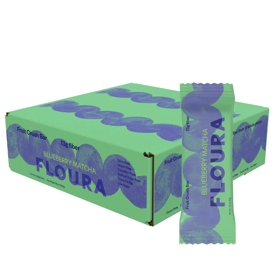 A green and purple FLOURA &amp; CO box labeled &quot;BLUEBERRY MATCHA FLOURS FIBERCRUSH BAR 2&quot; sits behind a matching fruit bar. The packaging highlights 13g prebiotic fiber, dietary benefits, and its value as a gut health snack.