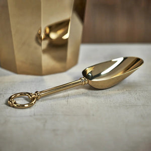 The ALESSIA GOLD ICE SCOOP by ZODAX gleams with its shiny gold finish and features a rounded handle measuring 10 inches. The handle ends in a circular ring, adding an elegant touch to the design, as it lies on a plain white background.