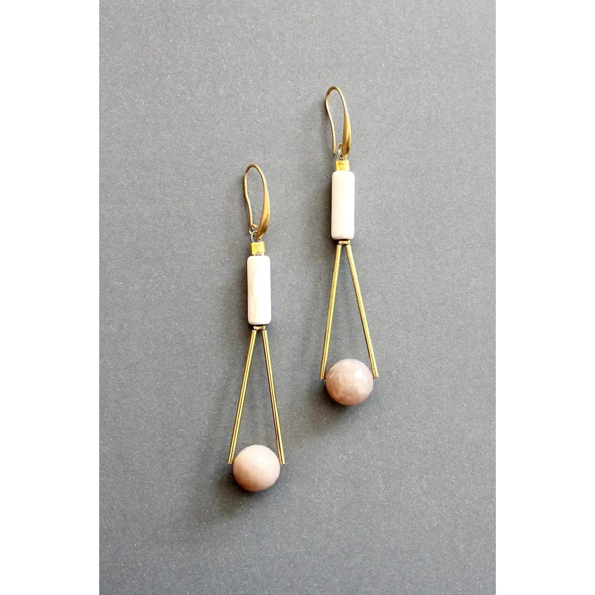 The DAVID AUBREY - PEACH MOONSTONE GEOMETRIC EARRINGS by DAVID AUBREY feature a sleek 18k gold plated triangular wire design, adorned with a white magnesite cylindrical bead at the top and a round peach moonstone bead at the bottom, showcased against a gray background.