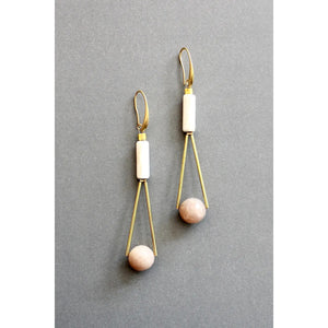 The DAVID AUBREY - PEACH MOONSTONE GEOMETRIC EARRINGS by DAVID AUBREY feature a sleek 18k gold plated triangular wire design, adorned with a white magnesite cylindrical bead at the top and a round peach moonstone bead at the bottom, showcased against a gray background.