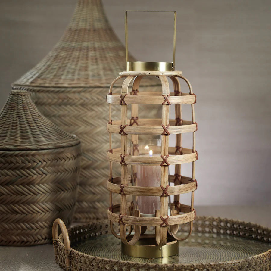 The Zodax Bamboo Lantern with Brass Trim is a decorative piece featuring a cylindrical glass holder encased in a crisscross bamboo lattice. Measuring 24.13 cm x 24.13 cm x 52.07 cm, it boasts metallic top and base elements with a convenient handle for easy carrying, seamlessly blending rustic and modern design elements.