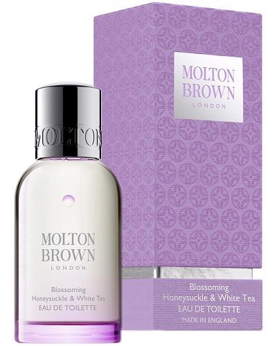 A clear bottle labeled &quot;Molton Brown&quot; containing &quot;Honeysuckle and White Tea Eau de Toilette,&quot; with a black cap, stands in front of its matching light purple box. The textured box features white labeling that states &quot;Molton Brown - Honeysuckle and White Tea Eau de Toilette,&quot; promising delightful floral fragrances.
